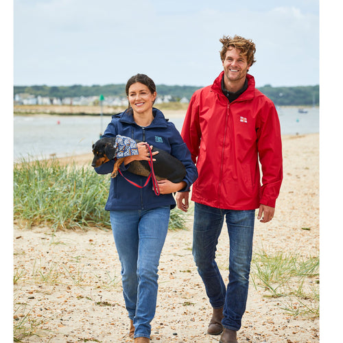 A man and woman walking on the beach with their dog. The man is wearing a red jacket with the RNLI flag logo on it.