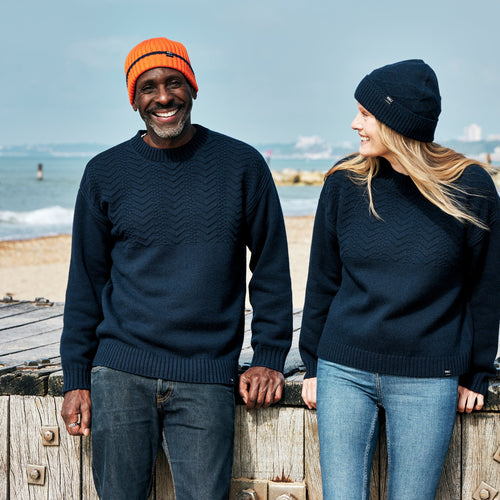 A man and a woman laughing together on the beach, both wearing RNLI + Finisterre Polperro Jumpers and matching beanies