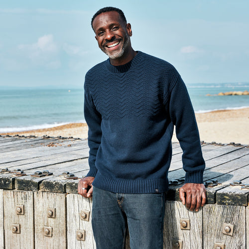 A man on the beach wearing the Men's RNLI + Finisterre Polperro Jumper in Navy and smiling widely