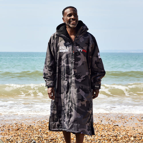 A man walking on the shoreline of a beach wearing a grey and black camouflage print dryrobe with the RNLI logo on the front.