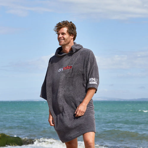 A man on a beach with the sea behind him wearing a grey RNLI dryrobe® Organic Towel Robe.