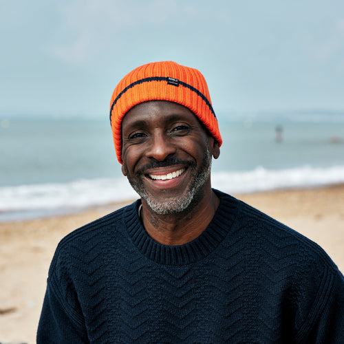 A man on a beach with the sea behind him, wearing an RNLI + Finisterre Fisherman Beanie in a bright orange colour. The beanie has a black stripe around the cuff and an embroidered logo.