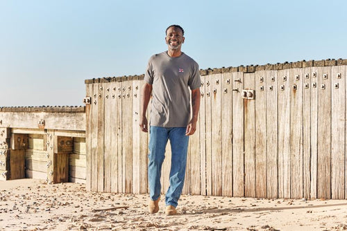 A person wearing a charcoal t-shirt with an embroidered flag on the chest, standing in front of a wooden structure.