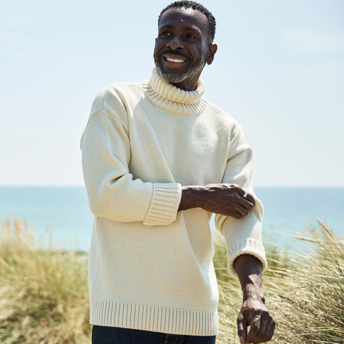 A man at the beach wearing a cream knitted jumper with a roll neck and rolled cuffs.