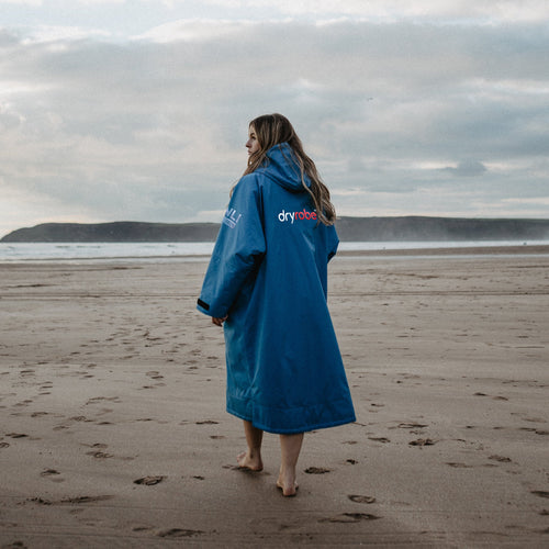 A woman walking on a stormy beach wearing a light blue dryrobe with long sleeves and a hood.