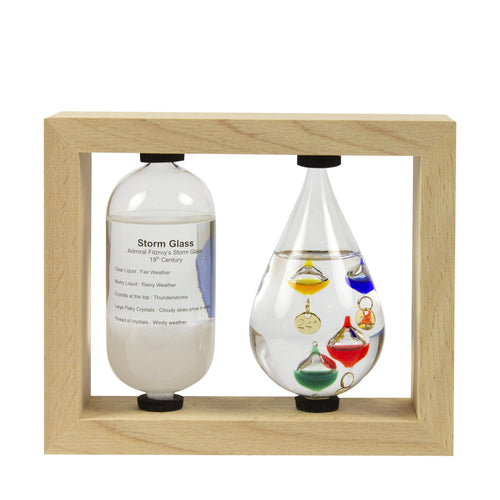 A Galileo thermometer and a storm bottle displayed in a wooden frame. The thermometer has colored bubbles inside a clear glass tube, and the storm bottle contains a mixture of liquids with colorful glass decorations representing weather indicators.