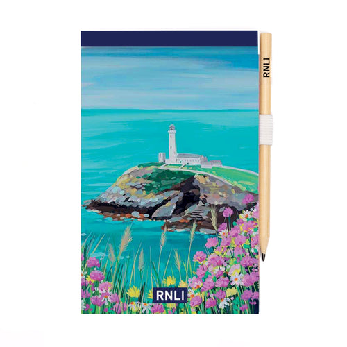 An A5 notepad with a painting of a lighthouse on top of a cliff on the cover. There are pink and yellow wildflowers in the foreground.