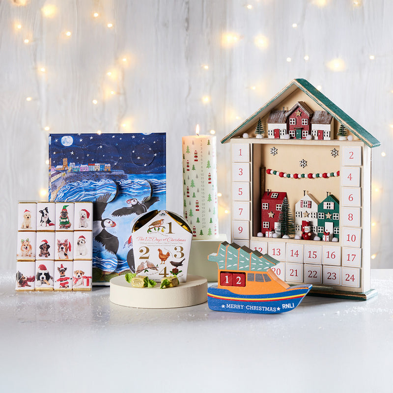  A collection of Christmas advent and holiday-themed products arranged on a white surface against a softly blurred white background with warm string lights. The set includes a large house-shaped wooden advent calendar with numbered doors, a smaller dog-themed advent calendar, a colourful RNLI lifeboat-shaped advent calendar.. 