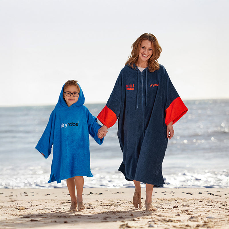  Adult and child on a beach. Adult is wearing a navy and orange RNLI and Dryrobe branded towel robe. Child is wearing Blue Dryrobe towel robe. 