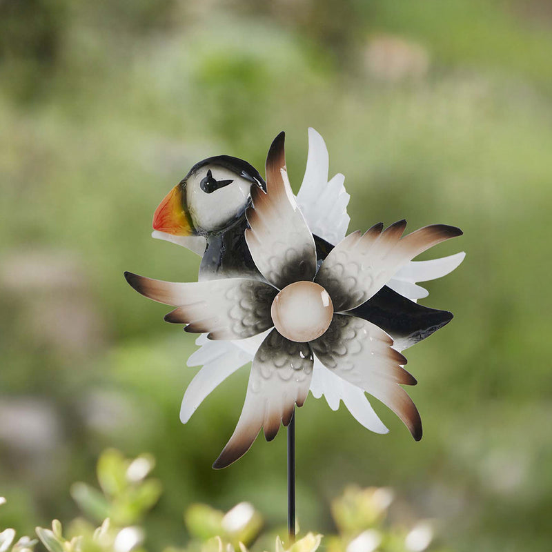  A decorative metal puffin garden wind spinner. The spinner is made up of puffin wings. It is on the top of a black garden stake. 