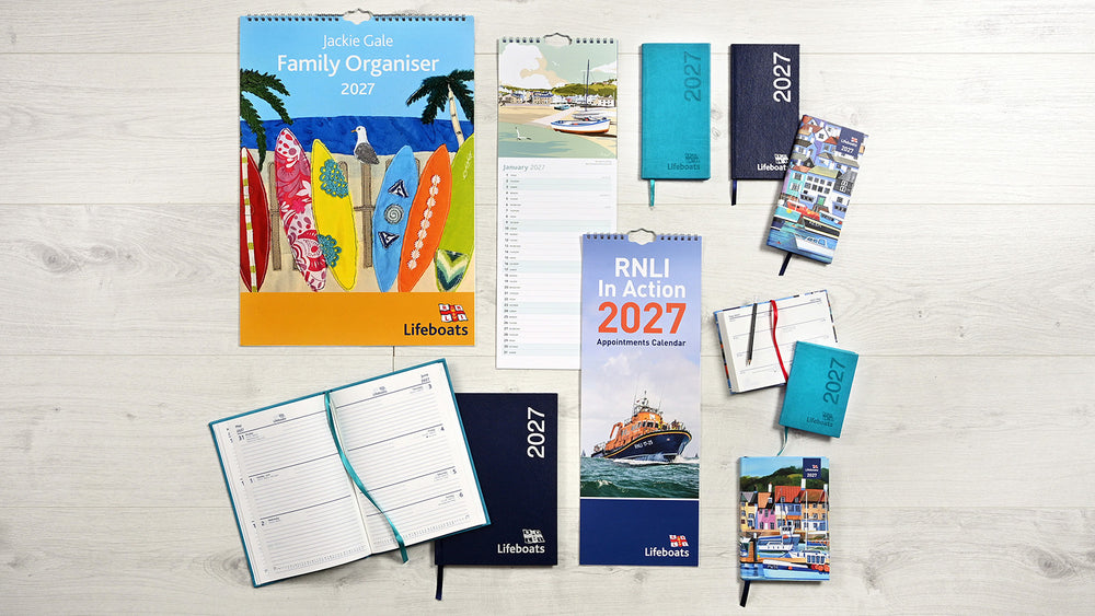  A flat-lay arrangement of multiple RNLI Lifeboats branded stationery and organiser products on a light wood background, including a large 2027 family organiser, a monthly calendar, notebooks, a planner, and other paper goods. 
