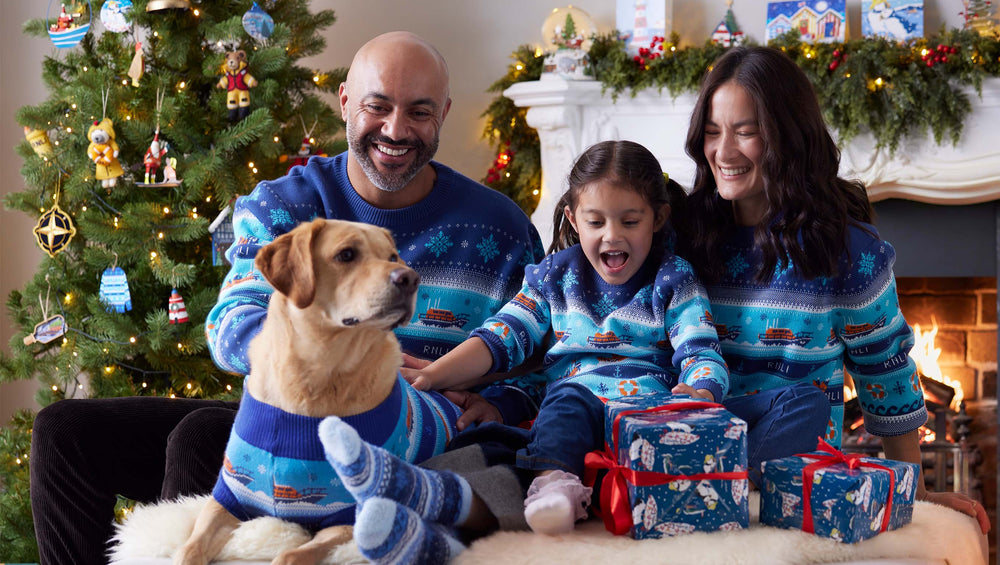  A family of four and their dog wearing matching blue RNLI Christmas jumpers and socks, gathered in a cosy living room with a lit Christmas tree, fireplace, and wrapped presents. 