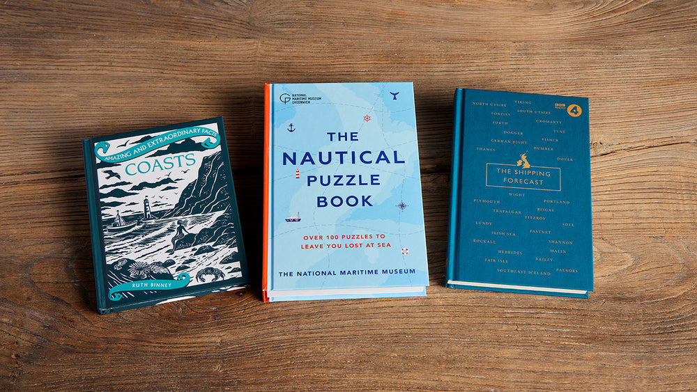  Three puzzle books arranged side by side on a wooden tabletop: 'Coasts' by Ruth Binney, 'The Nautical Puzzle Book' by The National Maritime Museum, and 'The Shipping Forecast' by Nic Compton. 