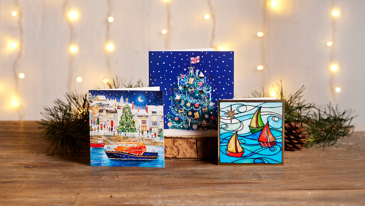 Three Christmas-themed cards displayed on a wooden surface with warm string lights and pine branches in the background. The left print shows a snowy village scene with an RNLI lifeboat, the center print shows a decorated Christmas tree with an RNLI flag on top on a blue starry background, and the right print shows stylized sailboats on water.