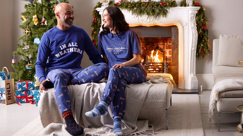  A man and woman sit in a cosy living room that's decorated for Christmas, with a Christmas tree, presents and open fire behind them. They are dressed in matching blue RNLI pyjamas, decorated with lifeboats. The man is wearing a sweater that reads 'in all weathers'. 