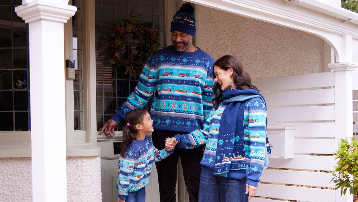 A smiling family of three stand on their front porch wearing matching RNLI Christmas jumpers and accessories. The striped, blue jumpers feature RNLI lifeboats, life rings and snowflakes.