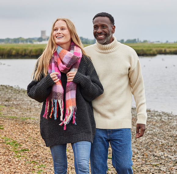  Two people standing by a body of water, one wearing a colourful scarf and a dark grey jumper, the other in a beige jumper. 