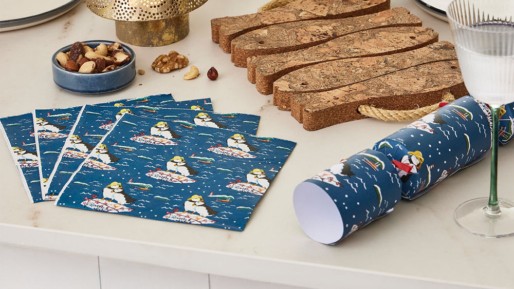  Roll of blue wrapping paper with an RNLI puffin design on a white surface with matching Christmas crackers and nuts and a fish shaped trivet. 