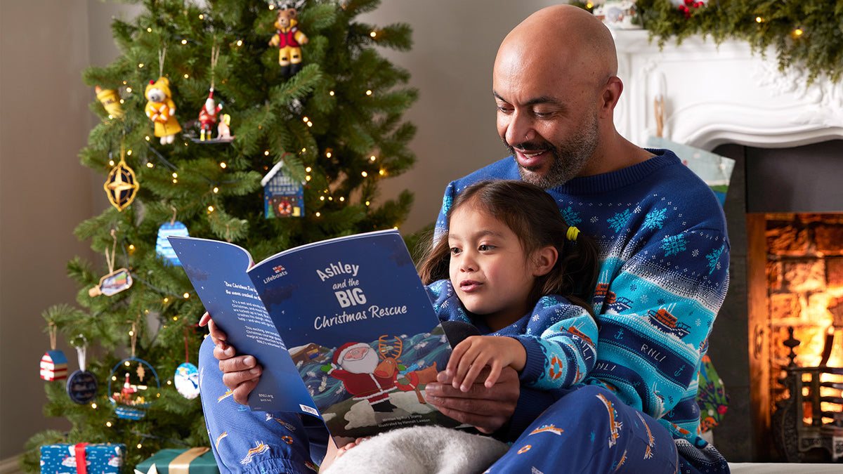  An adult and child wearing matching blue RNLI Christmas jumpers sit together in front of a fireplace, reading a blue personalised book titled 'Ashley and the Big Christmas Rescue' next to a decorated Christmas tree. 
