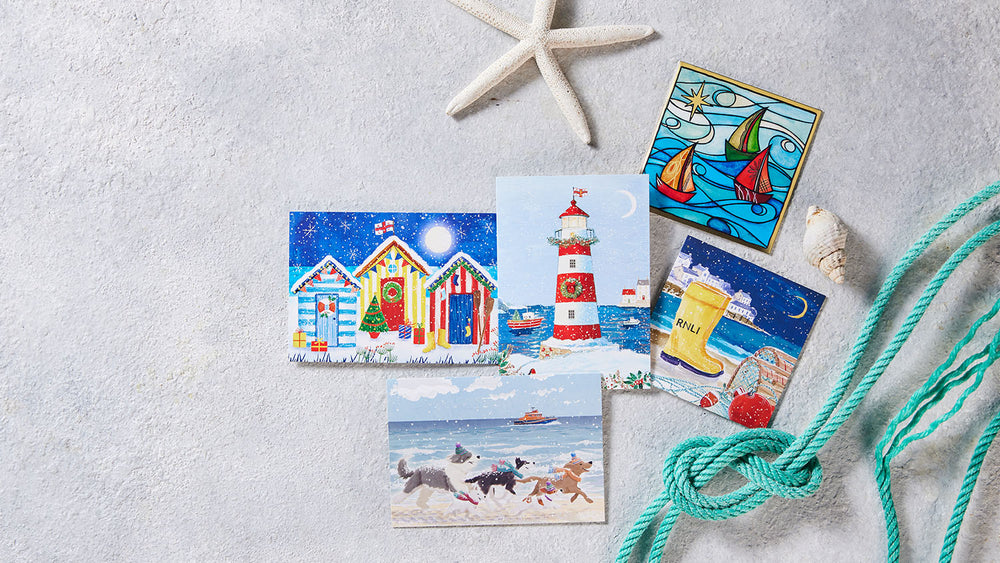  A set of illustrated beach and RNLI-themed Chritsmas cards arranged on a light textured surface with nautical props, including a starfish, shell, turquoise rope, and a lighthouse and beach scene card. 
