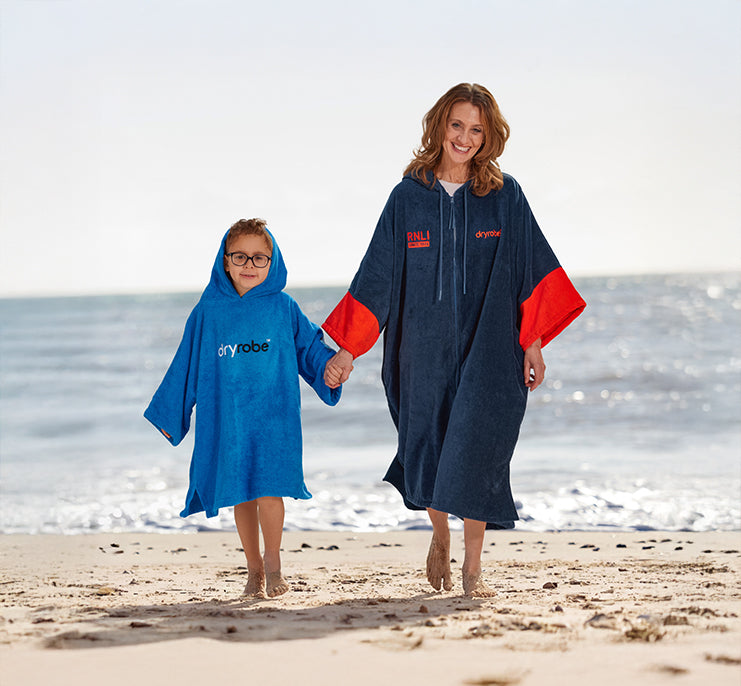  Two people wearing RNLI x Dryrobe® blue and orange ponchos on a beach with ocean in the background 