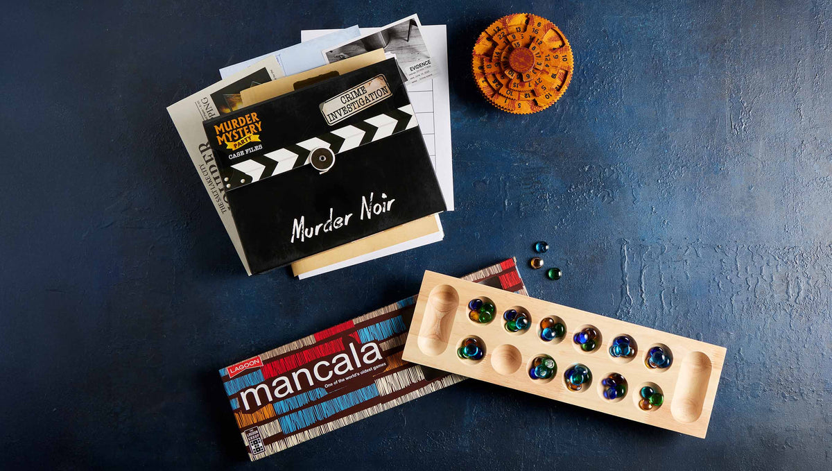 A styled flat-lay of a Murder Noir mystery game set on a dark blue textured background, including a black clapperboard-style box, a Mancala game board with colourful marbles, and a small round wooden puzzle.