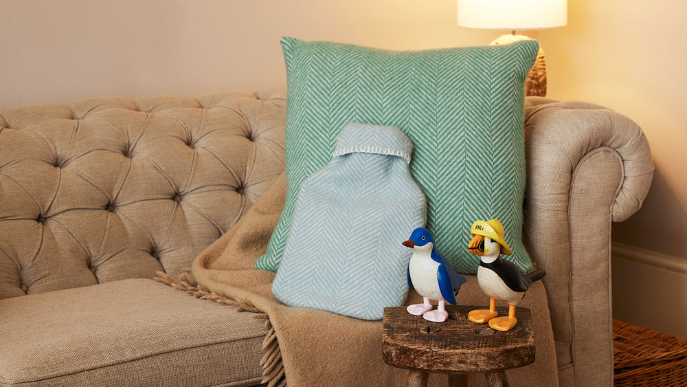  A styled living room scene featuring a beige chesterfield style sofa with a large mint green herringbone cushion and a light blue herringbone design fabric hot water bottle. Two small decorative penguin and puffin figurines sit on a rustic wooden block next to the sofa, with a warm lamp glowing in the background. 