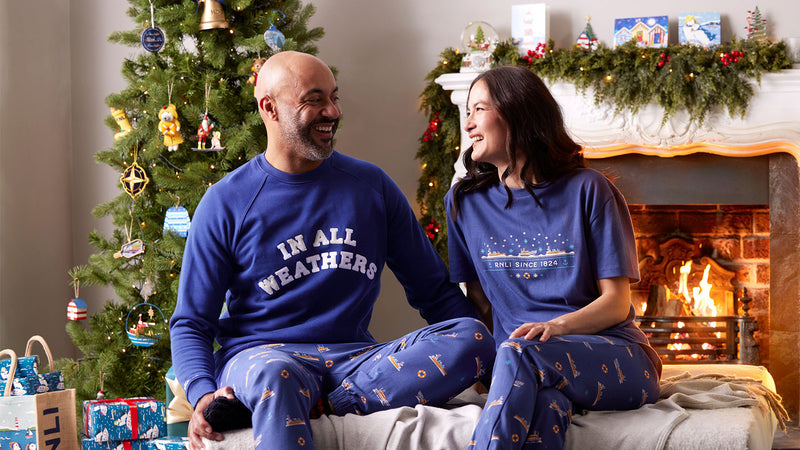 A man and a woman sitting together in front of a fireplace wearing matching blue Christmas lounge trousers. The man's sweatshirt reads 'IN ALL WEATHERS' and the woman's T-shirt reads 'RNLI SINCE 1824'. 