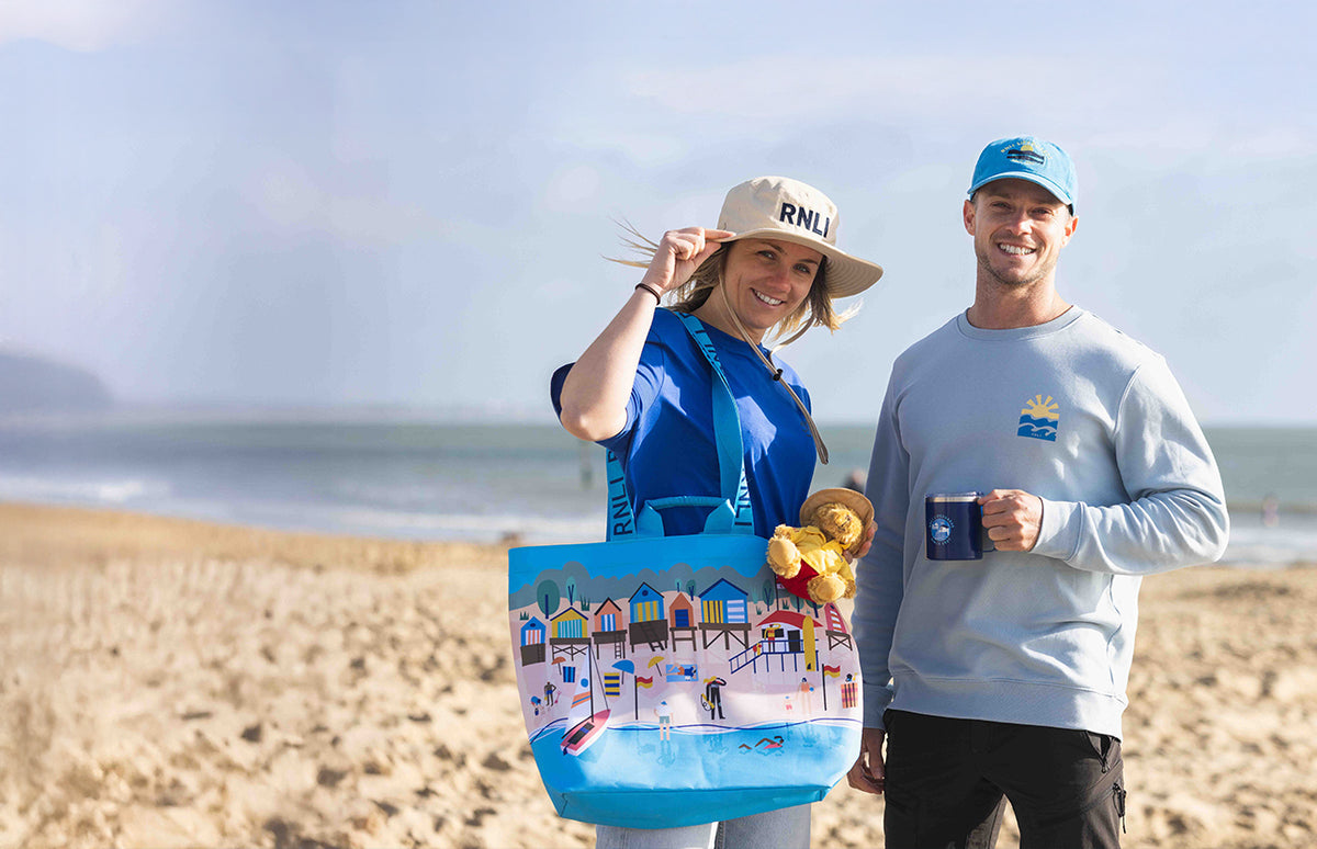  Two RNLI Lifeguards on a beach holding a crew member; Jonty Storey designed beach bag bag and a Lifeguards mug, with a clear sky in the background. Seen wearing some of the Lifeguards 25th Anniversary clothing and accessories. 