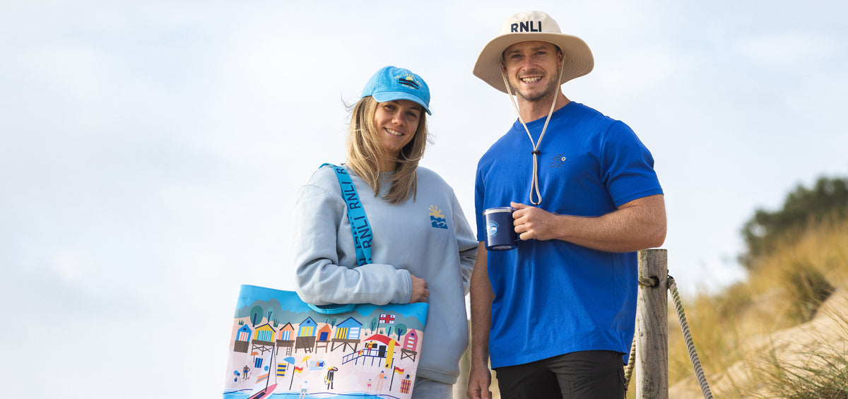  Two RNLI lifeguards standing outdoors with a scenic background, sporting clothing and accessories from the lifeguards 25th anniversary range 