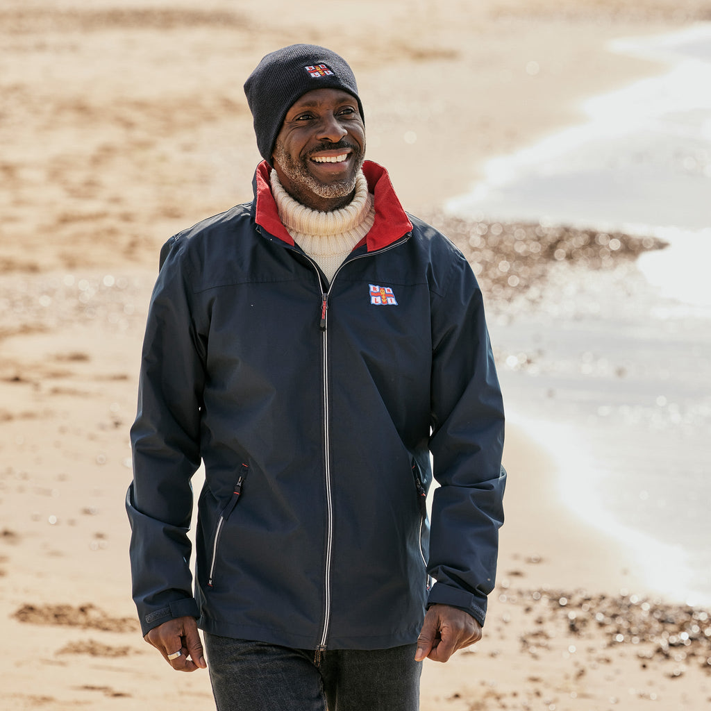 A man walking on the beach wearing a navy RNLI jacket and beanie hat.