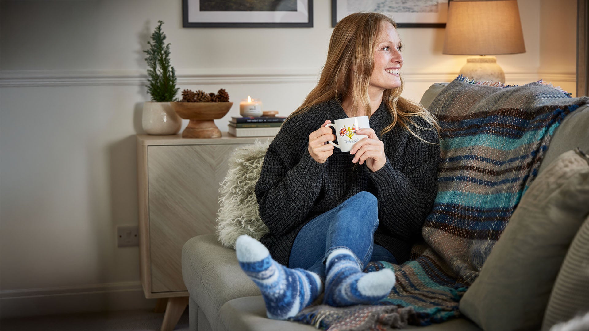 Woman wearing a dark grey jumper and light blue and dark blue fluffy socks sitting on a sofa with a blanket holding an RNLI  mug in a cosy living room.