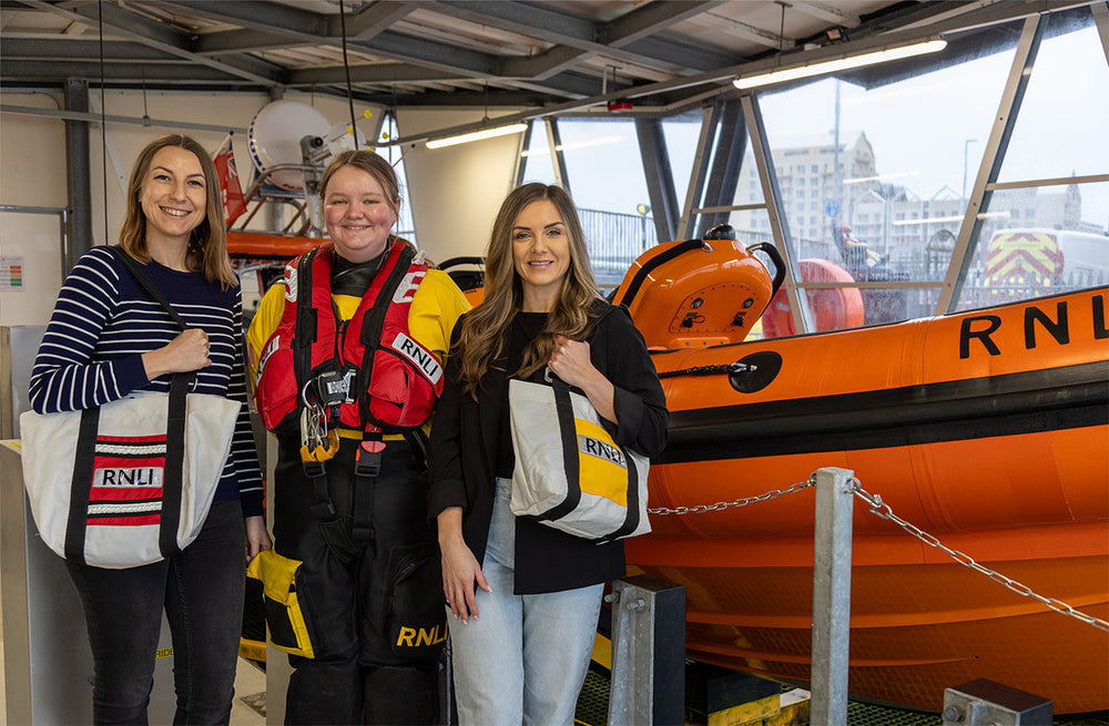  Three models standing in front of an orange RNLI lifeboat, one model is a crew member in their full crew kit, Two other models are sporting the RNLI repurposed ALB and ILB repurposed lifejacket tote bags. 
