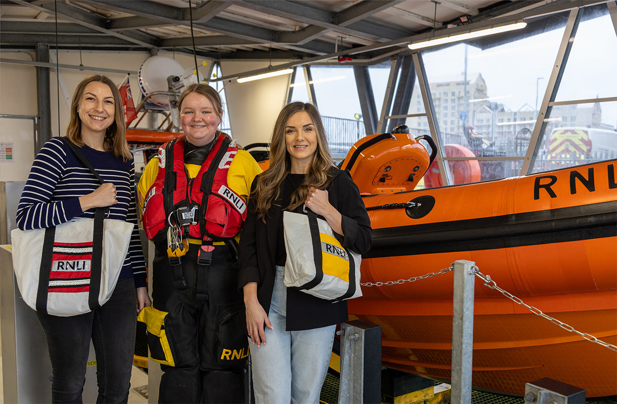  Three models standing in front of an orange RNLI lifeboat, one model is a crew member in their full crew kit, Two other models are sporting the RNLI repurposed ALB and ILB repurposed lifejacket tote bags. 