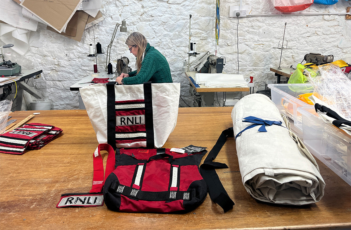  RNLI gear including a life jacket and bag on a table with a person working on sewing the jackets together to make the bag in the background. 
