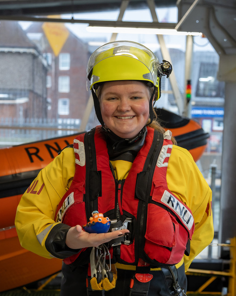  RNLI Crew member holding a toy lifeboat 'WOW Toys Lifeboat Lily' with real lifeboat in the background. 