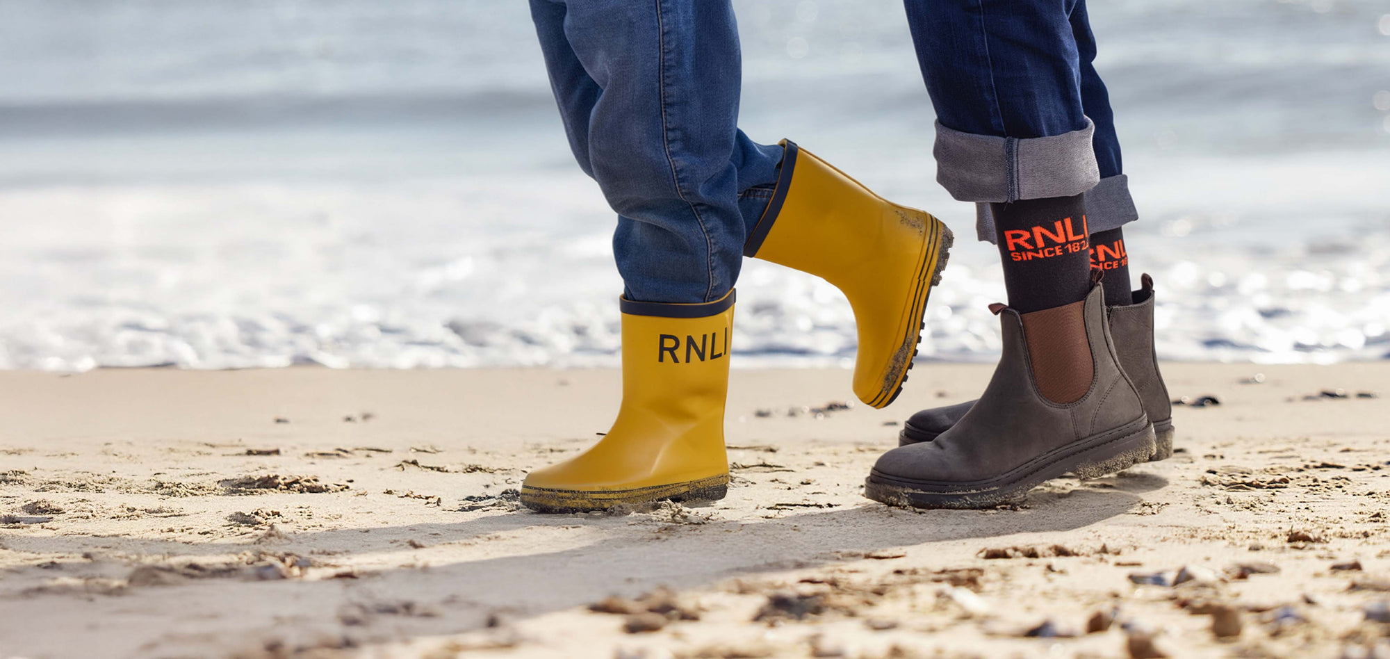 Two pairs of feet wearing yellow RNLI wellies and RNLI Since 1824 socks on a beach.