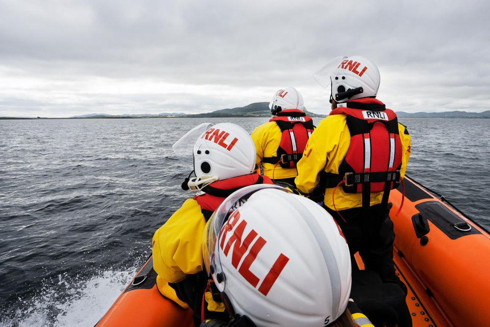  RNLI Lifeboat crew wearing yellow and red crew kit looking out towards the water from their boat, their white and red RNLI helmets facing the camera. 