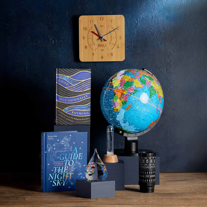  A styled tabletop arrangement featuring a colourful globe on a stand, a Bamboo wall clock, books including 'A Guide to the Night Sky', a Storm Glass, and a Galileo thermometer set against a dark blue wall. 
