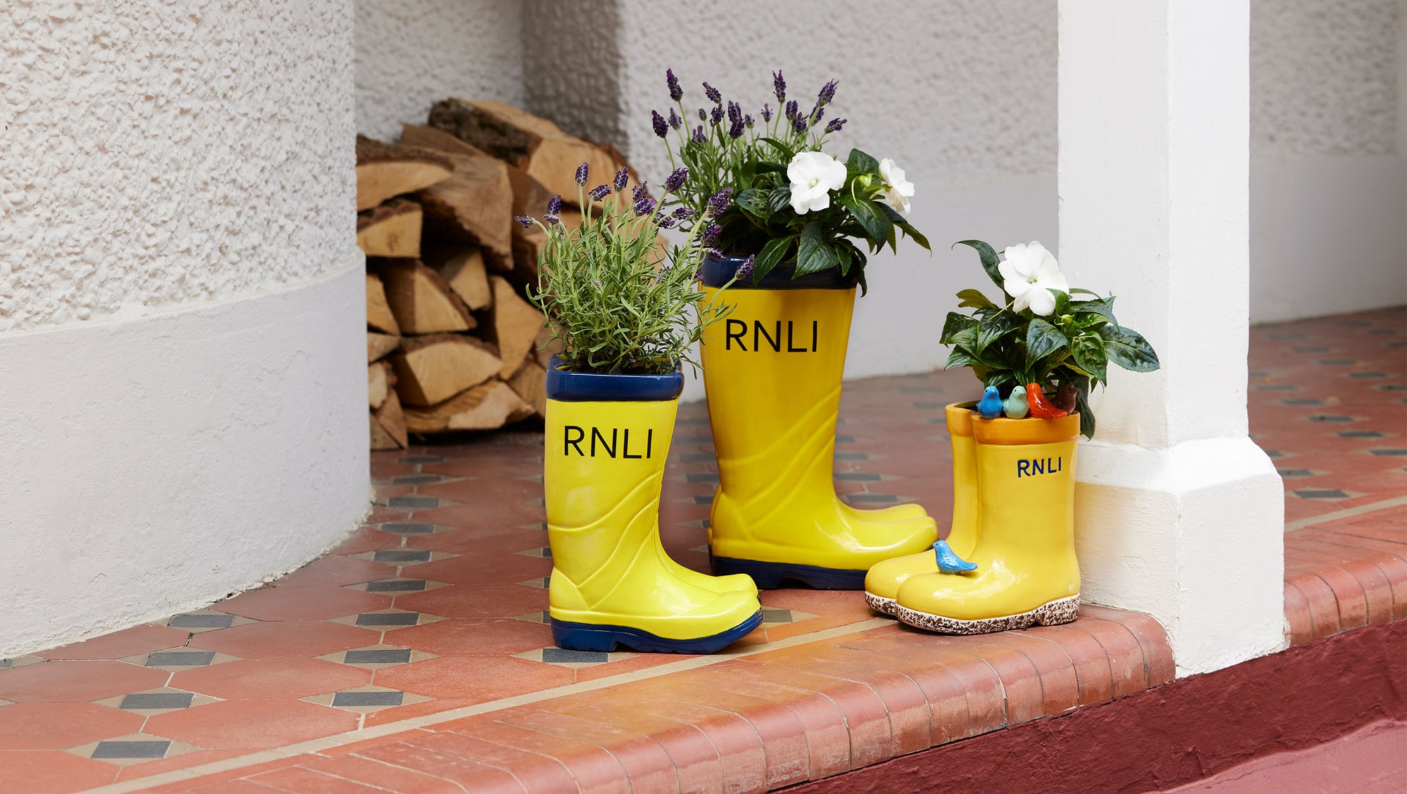 Three bright yellow rain boot planters with 'RNLI' printed on them,  with green foliage and white flowers on a terracotta tiled porch step.