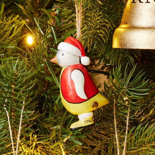 Red and yellow DCUK Penguin Hanging Decoration on a green Christmas tree with fairy lights and the edge of a gold bell.