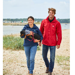 A man and woman walking on the beach with their dog. The man is wearing a red jacket with the RNLI flag logo on it.