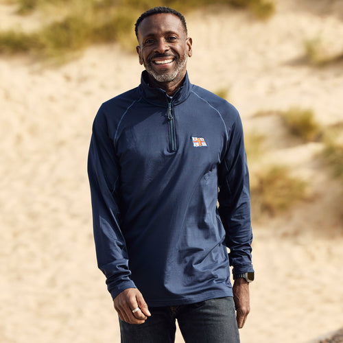 A man walking on a beach wearing a long-sleeved RNLI Sweater top in navy. The RNLI flag is printed on the left chest. 