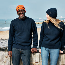 A man and a woman laughing together on the beach, both wearing RNLI + Finisterre Polperro Jumpers and matching beanies