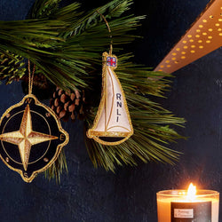 Two decorative hanging ornaments displayed among green pine branches against a dark navy background: a gold-toned compass ornament on the left and a sailboat ornament with a cream sail and vertical 'RNLI' text on the right. A lit candle is visible in the lower right corner.