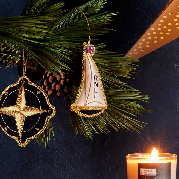 Two decorative hanging ornaments displayed among green pine branches against a dark navy background: a gold-toned compass ornament on the left and a sailboat ornament with a cream sail and vertical 'RNLI' text on the right. A lit candle is visible in the lower right corner.