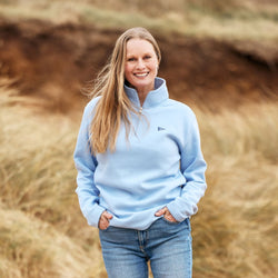 A model standing outdoors wearing a light blue half-zip sweatshirt with the RNLI burgee flag logo and blue jeans.