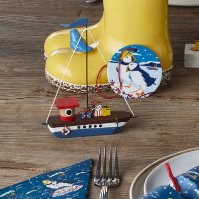 A small decorative sailboat ornament with a red roof, blue hull, and white mast, displayed on a rustic wooden table in front of a yellow welly planter and a round puffin-themed decoration.