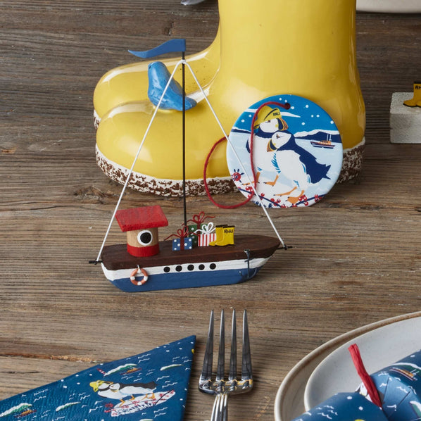 A small decorative sailboat ornament with a red roof, blue hull, and white mast, displayed on a rustic wooden table in front of a yellow welly planter and a round puffin-themed decoration.