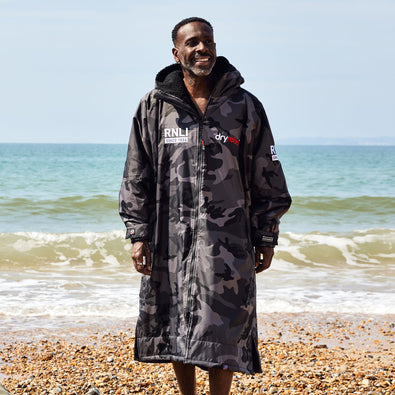 A man walking on the shoreline of a beach wearing a grey and black camouflage print dryrobe with the RNLI logo on the front.